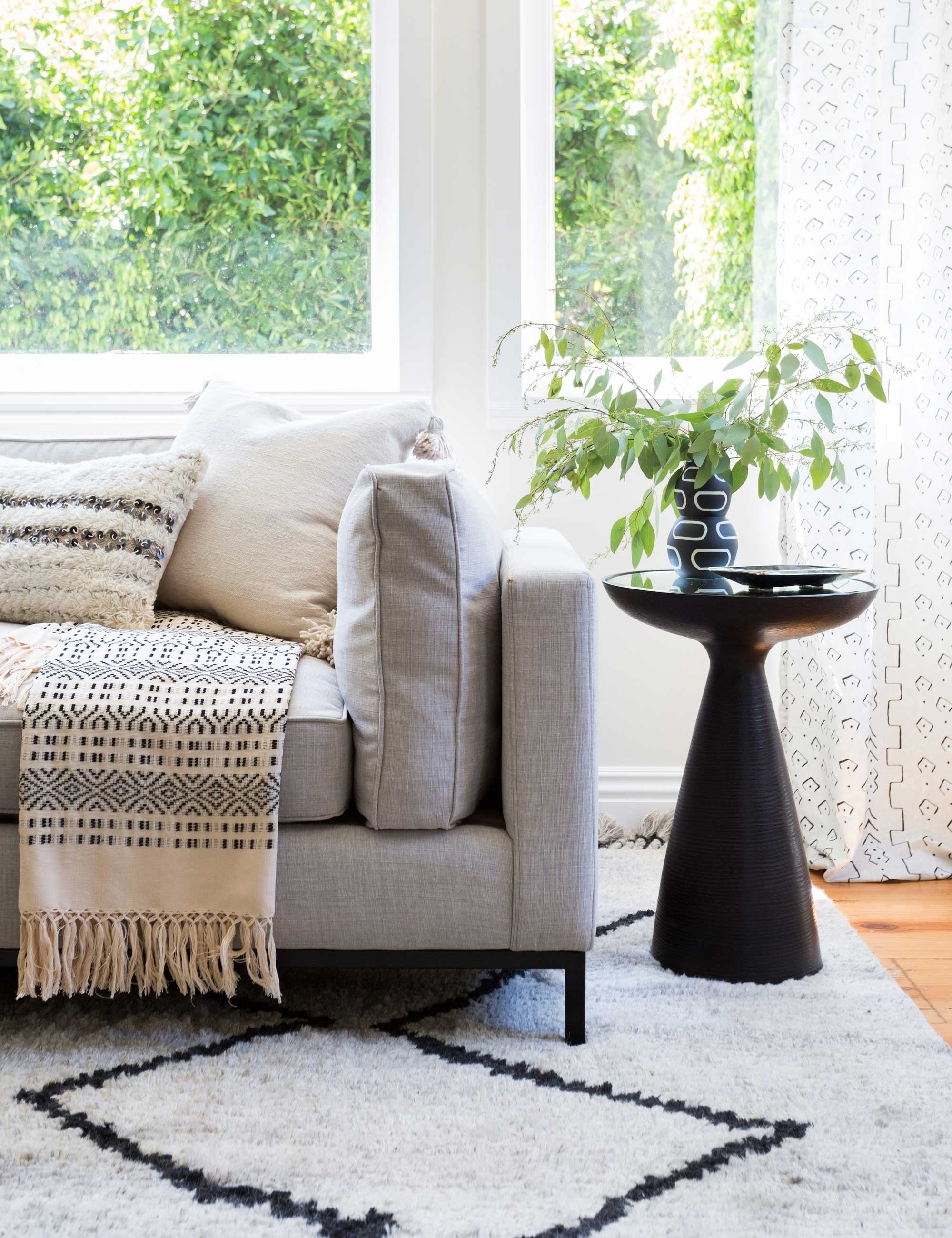 | The Bridget bronze pedestal side table with mirror ash glass top sits in a living room with a gray linen sofa, a plush patterned rug, and a geometric throw blanket